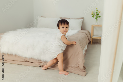 cute toddler girl standing near bed, smiling and looking at camera in apartment.
