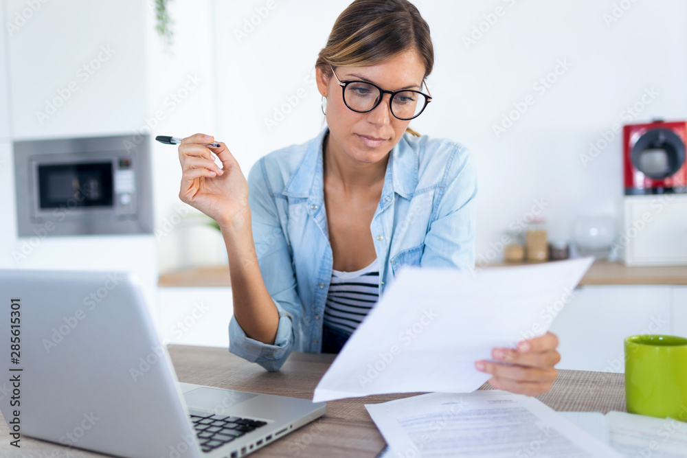 Pretty young woman working with laptop and documents in the kitchen at home. Pretty young woman working with laptop and documents in the kitchen at home.