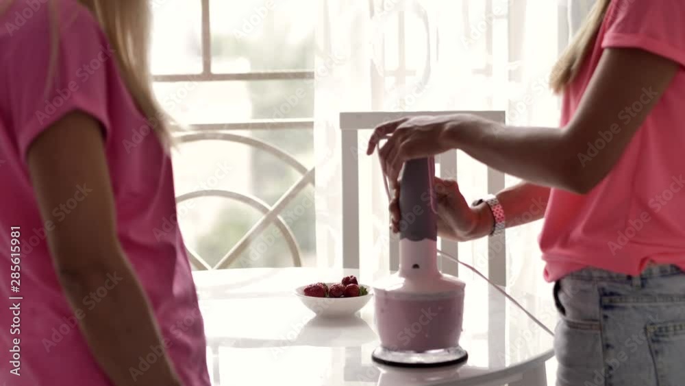 Two blondes, twin sisters, in pink clothes, cook something together, crush and mix a milkshake with a blender, on a table there is a plate with strawberries, natural light.