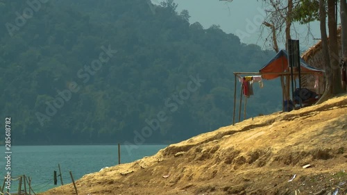 Close-up low-angle view of exposed aboriginal home ground slanting to the lake bank, native houses and structures, calm greenish water of Temenggor lake, and fogy Rainforest highland on the opposite photo