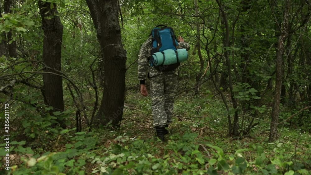 young handsome сaucasian man in a camouflage suit, cap and with a backpack, walks through the dense green forest past the camera, the average plan young handsome сaucasian man in a camouflage suit, cap and with a backpack, walks through the dense green forest past the camera, the average plan