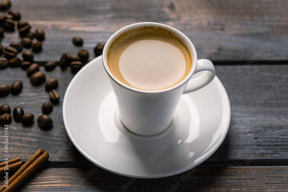 Cup of black coffee on a saucer with cinnamon on a dark wooden table. Coffee beans scattered on the table Cup of black coffee on a saucer with cinnamon on a dark wooden table. Coffee beans scattered on the table