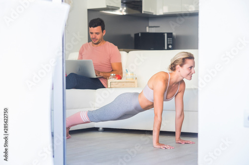 Fitness at home wellness actiity with young caucasian people couple - man working with technology laptop and blonde beautiful girl doing push and balance position to build perfect body