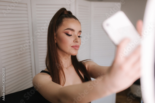 Young attractive model make a selfie after makeup in beauty studio. She use a ring lapm for photo. Background of white folding screen photo