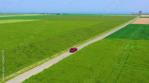 Aerial of a red car traveling through green fields of Normandy, France.