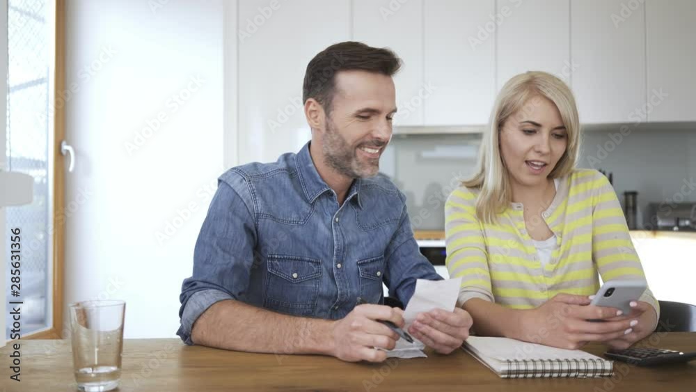Cheerful couple managing home finance with smartphone and bills then embracing Cheerful couple managing home finance with smartphone and bills then embracing