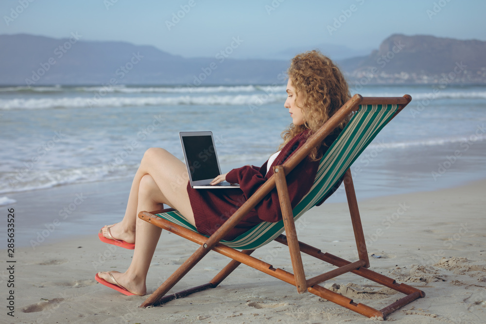 Woman using laptop while sitting on sun lounger at beach 
