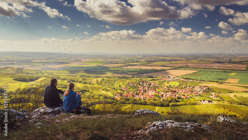 Two people (couple - man and woman) sitting on a mountain on a stone and looking into the valley on beautiful autumn landscape with village, vineyards and fields. Blue sky, white clouds. Palava, Czech photo