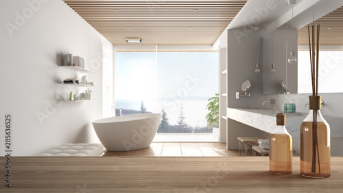 Wooden table top or shelf with aromatic sticks bottles over blurred modern minimalist luxury bathroom with bathtub and panoramic window  white architecture interior design