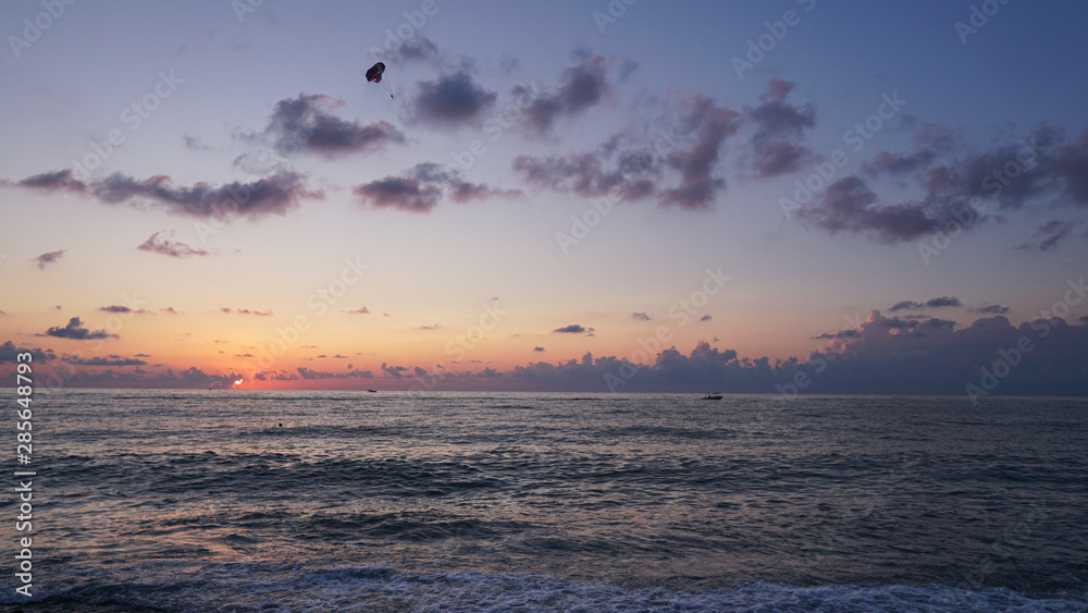 Parasailing in blue sea during orange evening sundown on horizon, beautiful scenery of tranquil seascape in summer evening twilights. Tourists on a parachute over the sea. Sunset paradise 