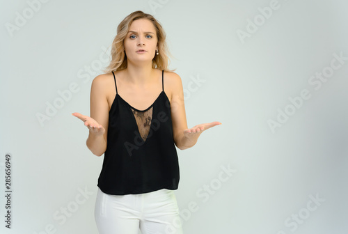 Photo studio portrait of a cute blonde young woman girl in a black blouse and white pants on a white background. He stands right in front of the camera, explains with emotion.