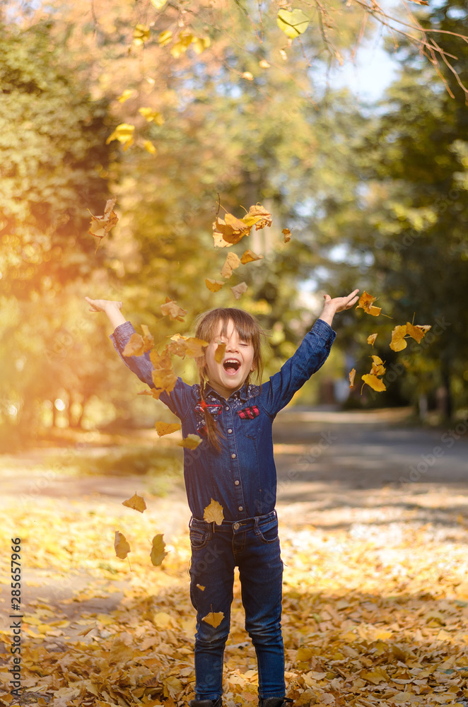 Beautiful little girl in denim shirt and jeans is sitting on yellow leaves and playing on warm and sunny autumn day. Happy childhood concept. Sun flare Beautiful little girl in denim shirt and jeans is sitting on yellow leaves and playing on warm and sunny autumn day. Happy childhood concept. Sun flare