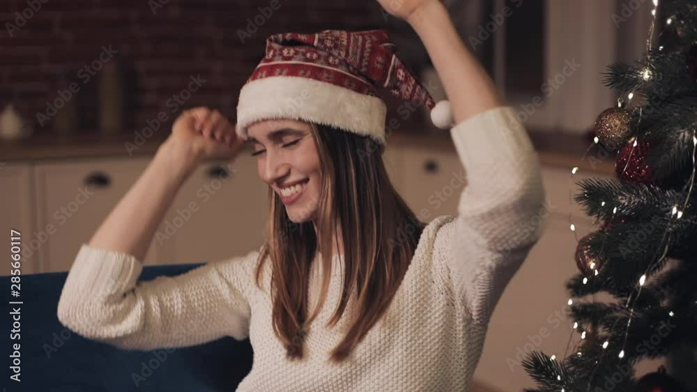 Young Attractive Girl Wearing Christmas Hat Having Fun Sitting on the Sofa with Smartphone in her hand Dances Rejoices Smiles and Laughs. Concept of Christmas holidays and New Year. Young Attractive Girl Wearing Christmas Hat Having Fun Sitting on the Sofa with Smartphone in her hand Dances Rejoices Smiles and Laughs. Concept of Christmas holidays and New Year.