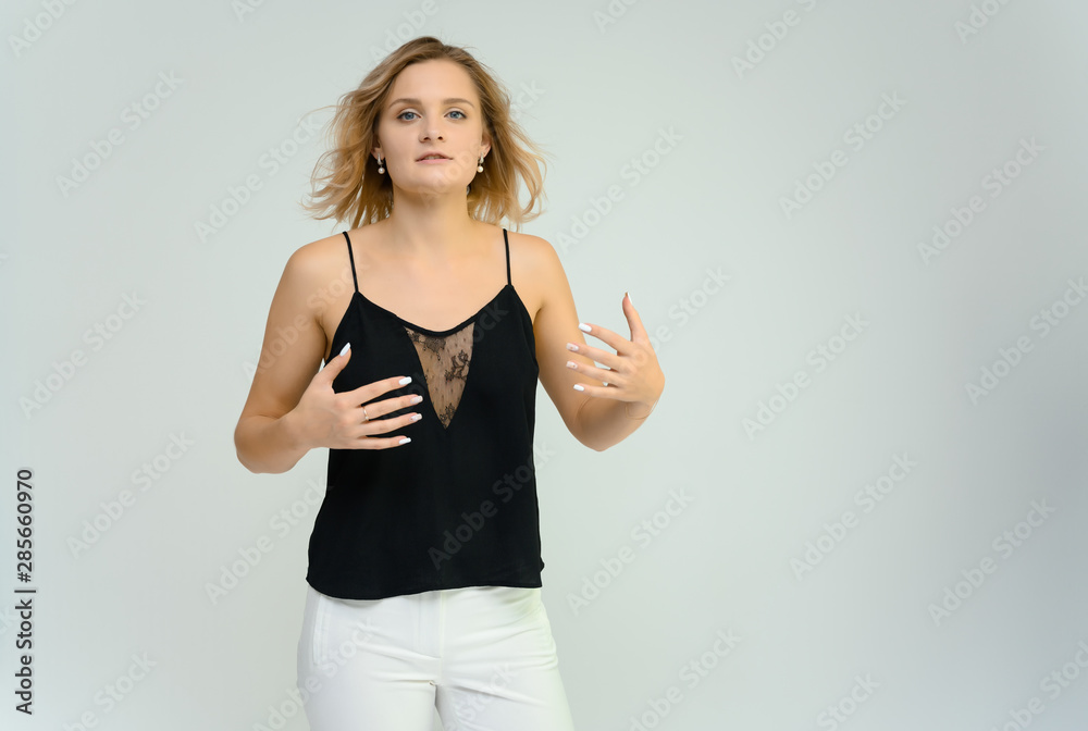 Photo studio portrait of a cute blonde young woman girl in a black blouse and white pants on a white background. He stands right in front of the camera, explains with emotion. Photo studio portrait of a cute blonde young woman girl in a black blouse and white pants on a white background. He stands right in front of the camera, explains with emotion.