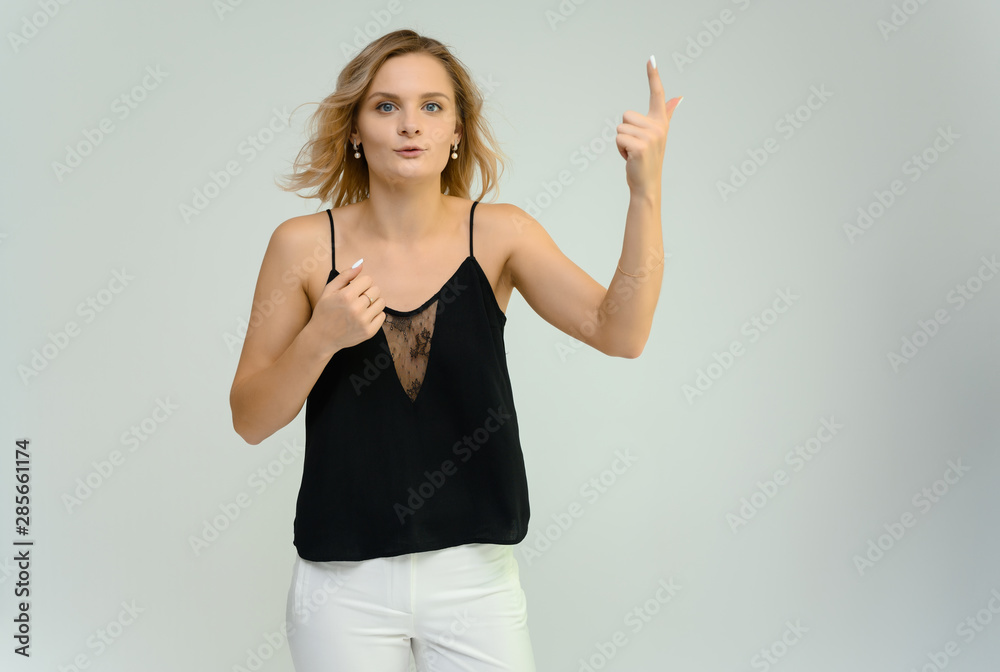 Fototapeta premium Photo studio portrait of a cute blonde young woman girl in a black blouse and white pants on a white background. He stands right in front of the camera, explains with emotion.