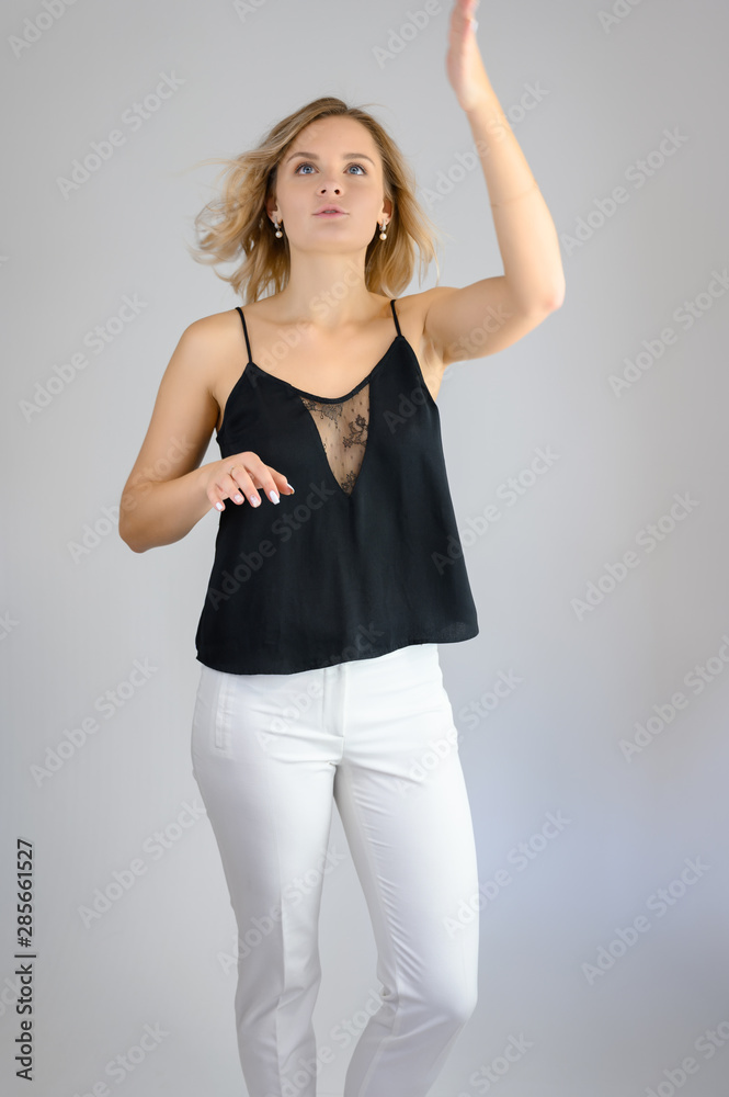 Full length studio portrait photo of a cute young blonde woman girl in a black blouse and white pants on a white background. He stands right in front of the camera, explains with emotion. Full length studio portrait photo of a cute young blonde woman girl in a black blouse and white pants on a white background. He stands right in front of the camera, explains with emotion.