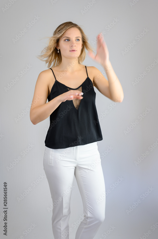 Full length studio portrait photo of a cute young blonde woman girl in a black blouse and white pants on a white background. He stands right in front of the camera, explains with emotion.
