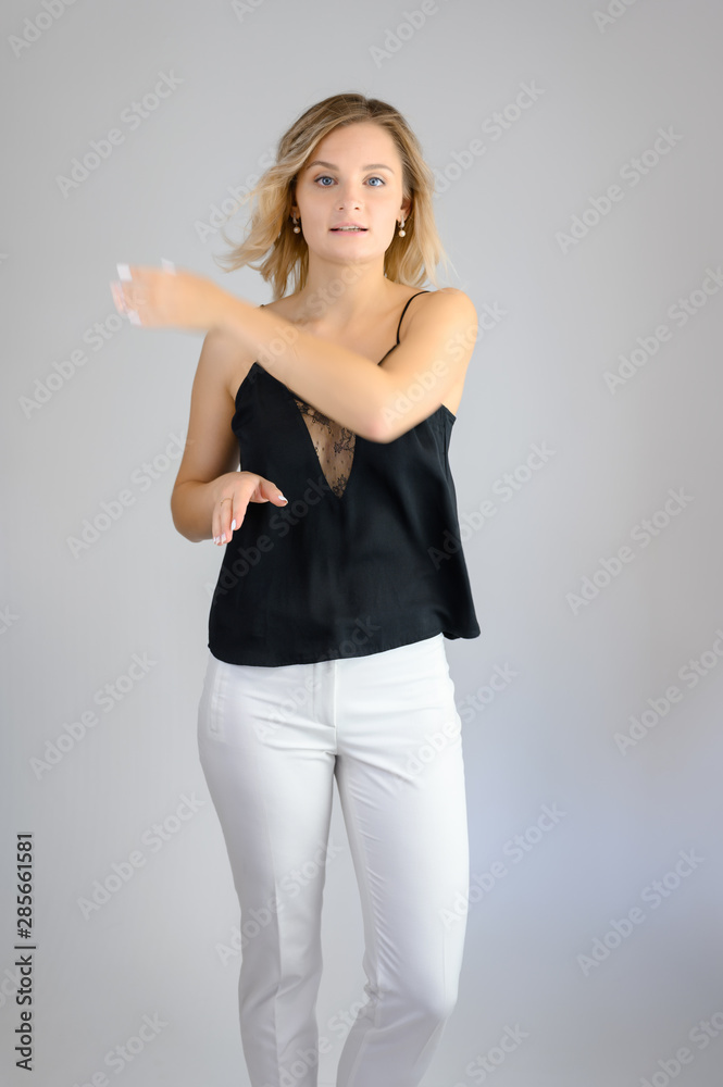 Full length studio portrait photo of a cute young blonde woman girl in a black blouse and white pants on a white background. He stands right in front of the camera, explains with emotion. Full length studio portrait photo of a cute young blonde woman girl in a black blouse and white pants on a white background. He stands right in front of the camera, explains with emotion.