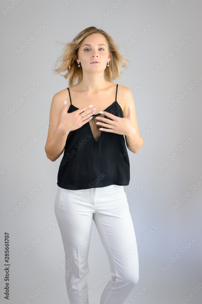 Full length studio portrait photo of a cute young blonde woman girl in a black blouse and white pants on a white background. He stands right in front of the camera, explains with emotion. Full length studio portrait photo of a cute young blonde woman girl in a black blouse and white pants on a white background. He stands right in front of the camera, explains with emotion.