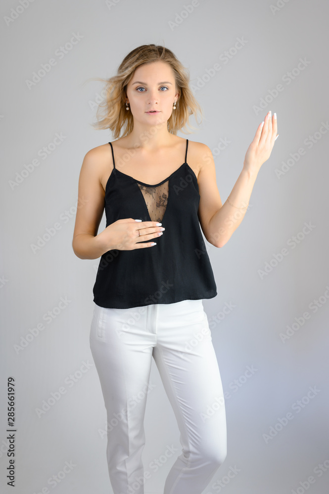 Full length studio portrait photo of a cute young blonde woman girl in a black blouse and white pants on a white background. He stands right in front of the camera, explains with emotion. Full length studio portrait photo of a cute young blonde woman girl in a black blouse and white pants on a white background. He stands right in front of the camera, explains with emotion.