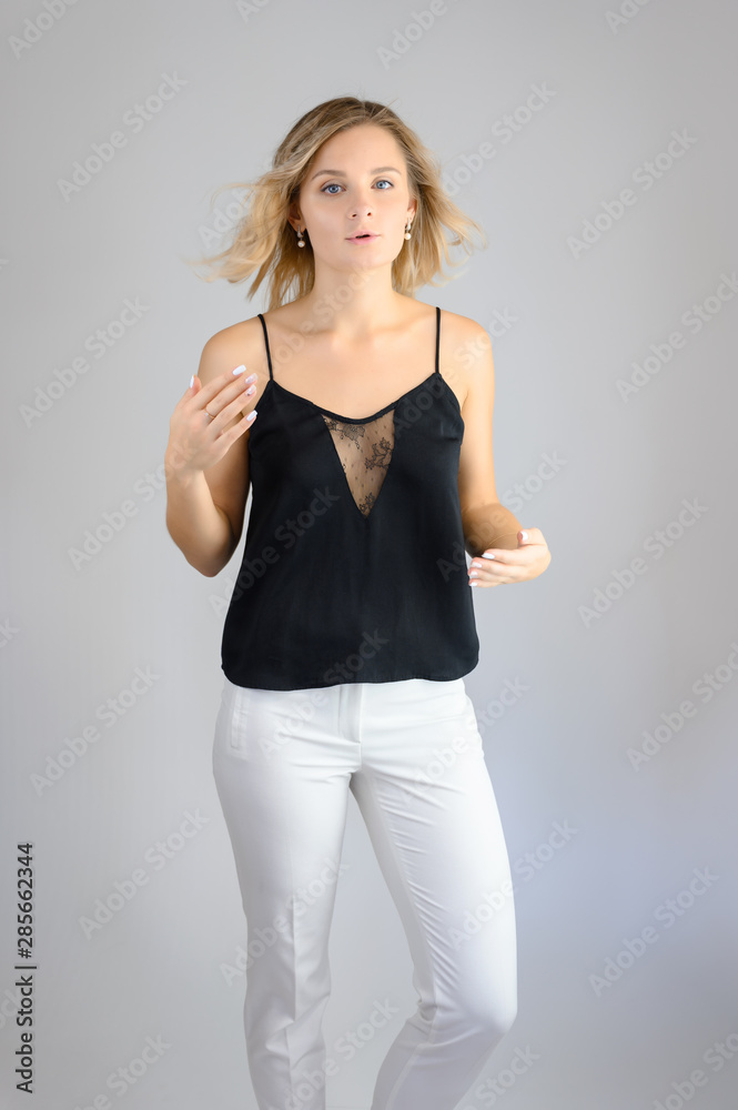 Full length studio portrait photo of a cute young blonde woman girl in a black blouse and white pants on a white background. He stands right in front of the camera, explains with emotion. Full length studio portrait photo of a cute young blonde woman girl in a black blouse and white pants on a white background. He stands right in front of the camera, explains with emotion.