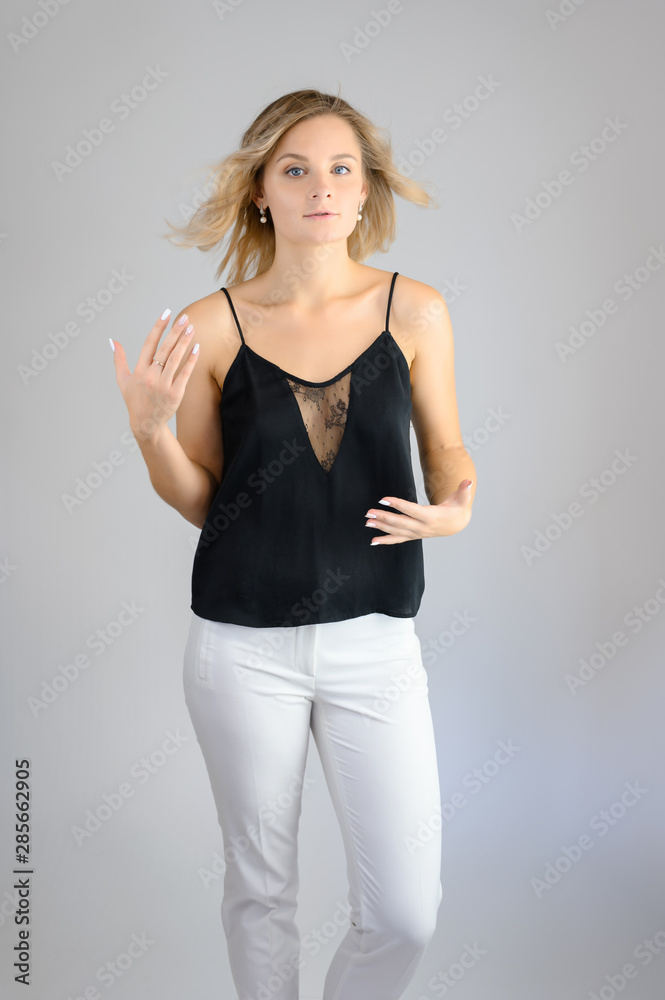 Full length studio portrait photo of a cute young blonde woman girl in a black blouse and white pants on a white background. He stands right in front of the camera, explains with emotion. Full length studio portrait photo of a cute young blonde woman girl in a black blouse and white pants on a white background. He stands right in front of the camera, explains with emotion.