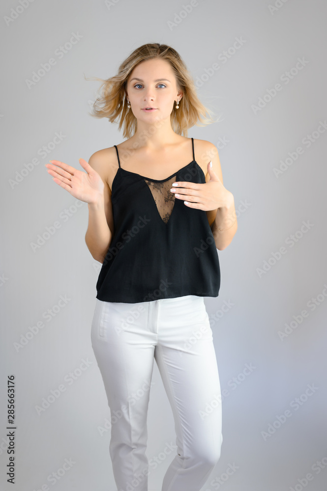 Full length studio portrait photo of a cute young blonde woman girl in a black blouse and white pants on a white background. He stands right in front of the camera, explains with emotion. Full length studio portrait photo of a cute young blonde woman girl in a black blouse and white pants on a white background. He stands right in front of the camera, explains with emotion.