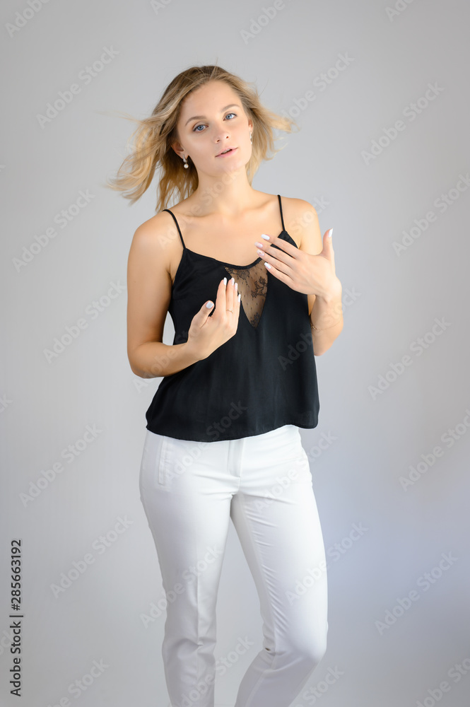 Full length studio portrait photo of a cute young blonde woman girl in a black blouse and white pants on a white background. He stands right in front of the camera, explains with emotion. Full length studio portrait photo of a cute young blonde woman girl in a black blouse and white pants on a white background. He stands right in front of the camera, explains with emotion.