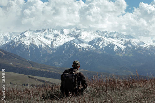 Man in camouflage sitting in dead grass in front of mountains