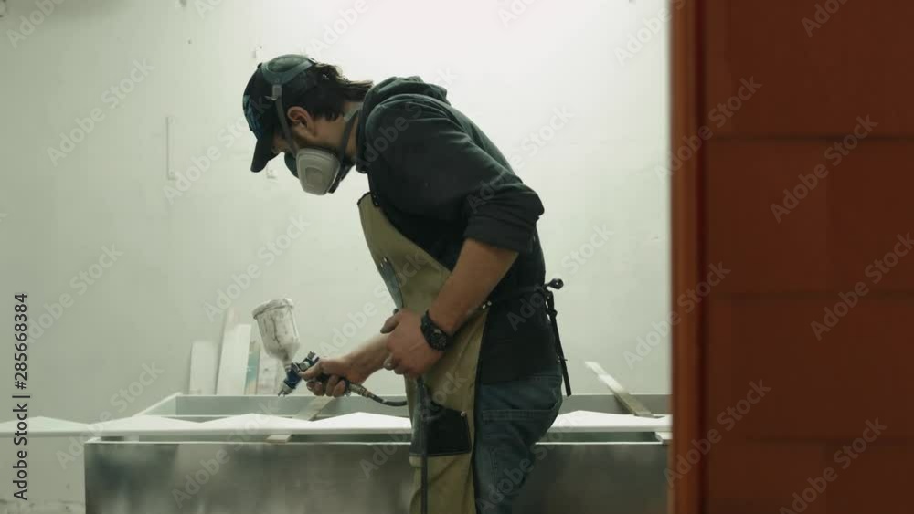 Man in respirator mask painting wooden planks at workshop. Craftsman modern furniture factory paint wood detail with spray gun, male professional housepainer