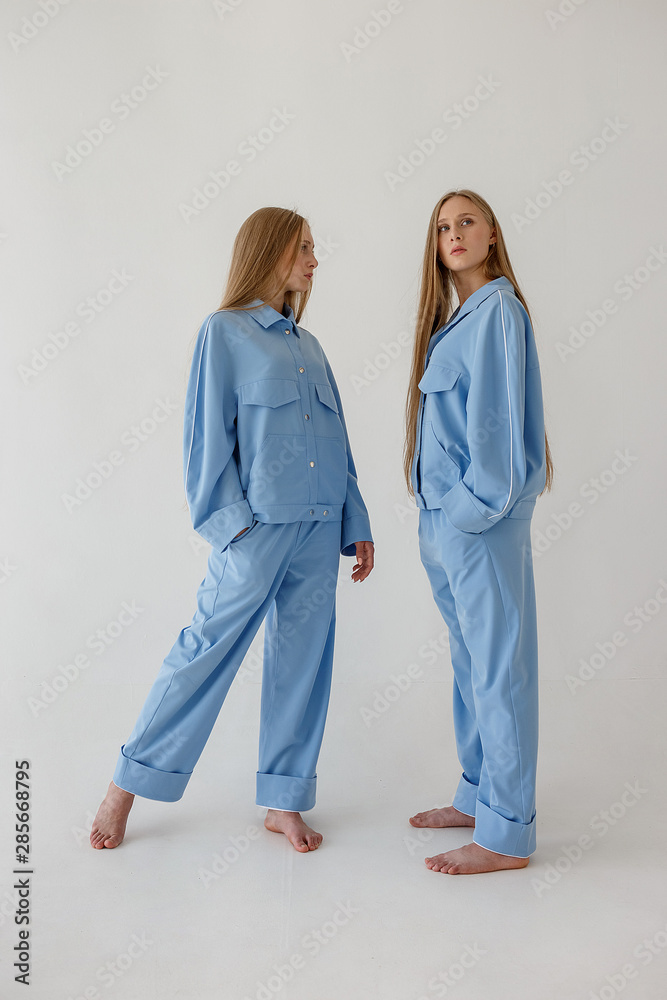 two pretty young twin sisters with long blond hair posing on white background in oversize clothes. Fashion photoshoot two pretty young twin sisters with long blond hair posing on white background in oversize clothes. Fashion photoshoot