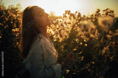 Happy young woman is walking among the flowers and grass on meadow at sunset.
