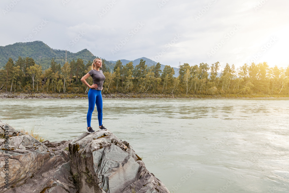 Blond beautiful strong girl standing on the stone cliff ground resting after sport outdoors in the mountains near the river. Workout in nature.