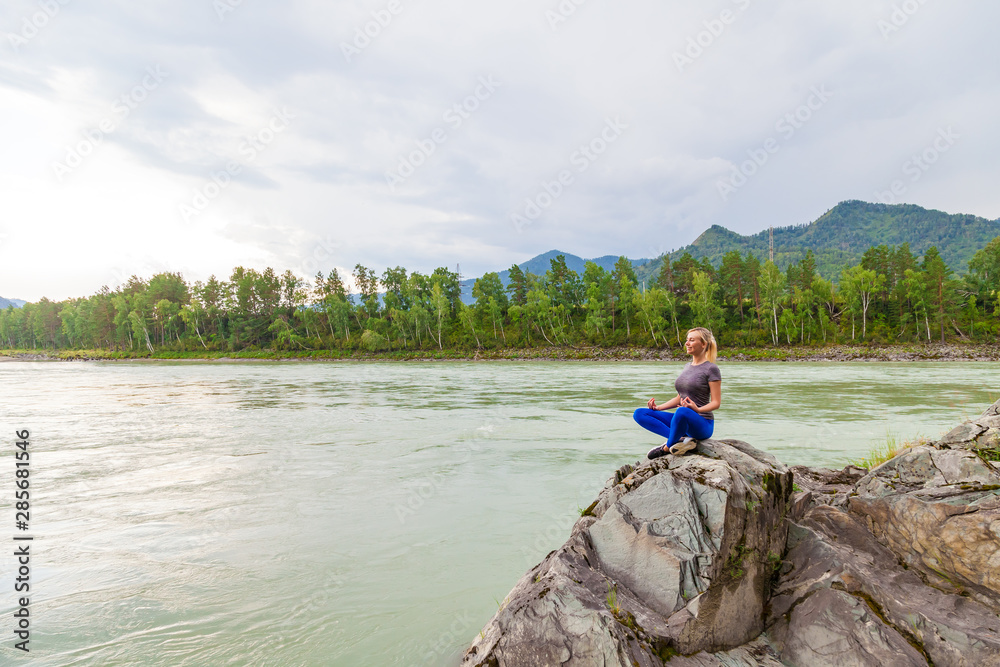 Blond beautiful girl sitting on the stone cliff ground resting after sport and doing yoga and meditation outdoors in the mountains near the river. Workout and relaxation in nature.