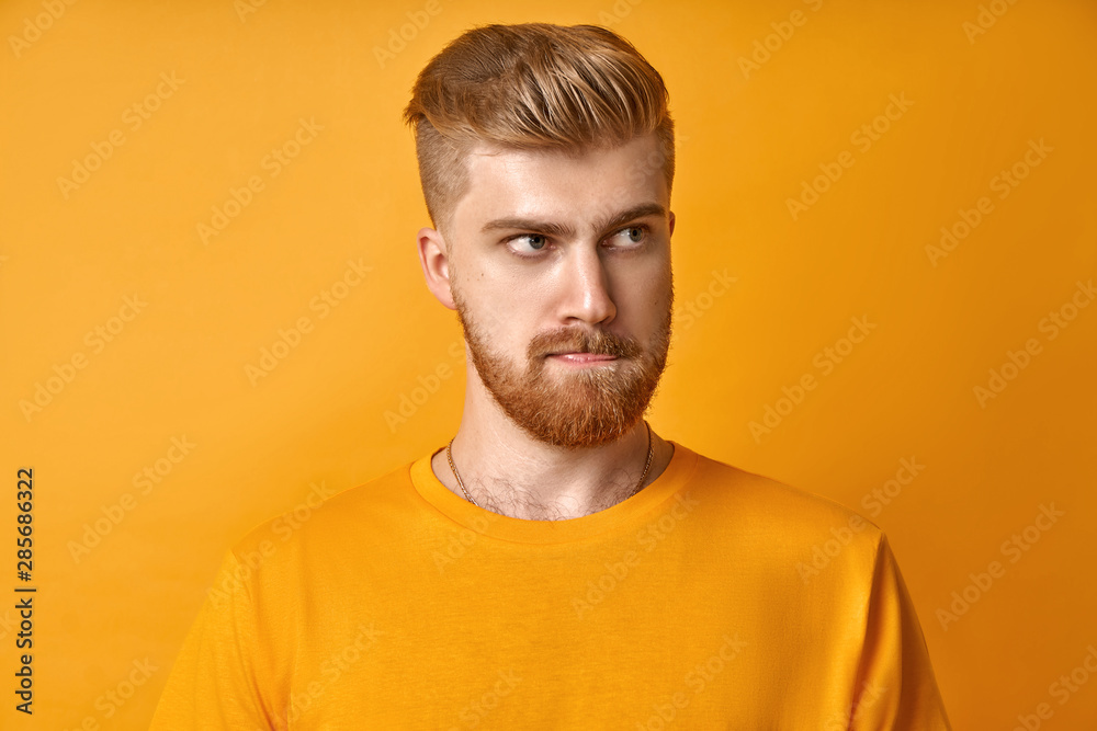 Obraz premium Puzzled handsome male with mustache and red beard, bites lower lip and looks curiously aside, thinks about something, dressed in casual yellow t shirt, stands against yellow background