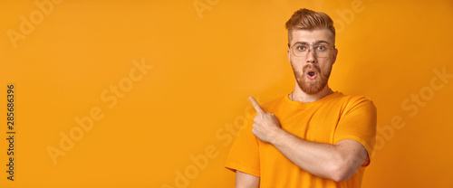 Surprised male hipster with trendy hairdo and red beard, keeps mouth opened, being stupefied as advertises something, wears yellow tshirt, indicates with fore finger at upper right corner. Omg concept