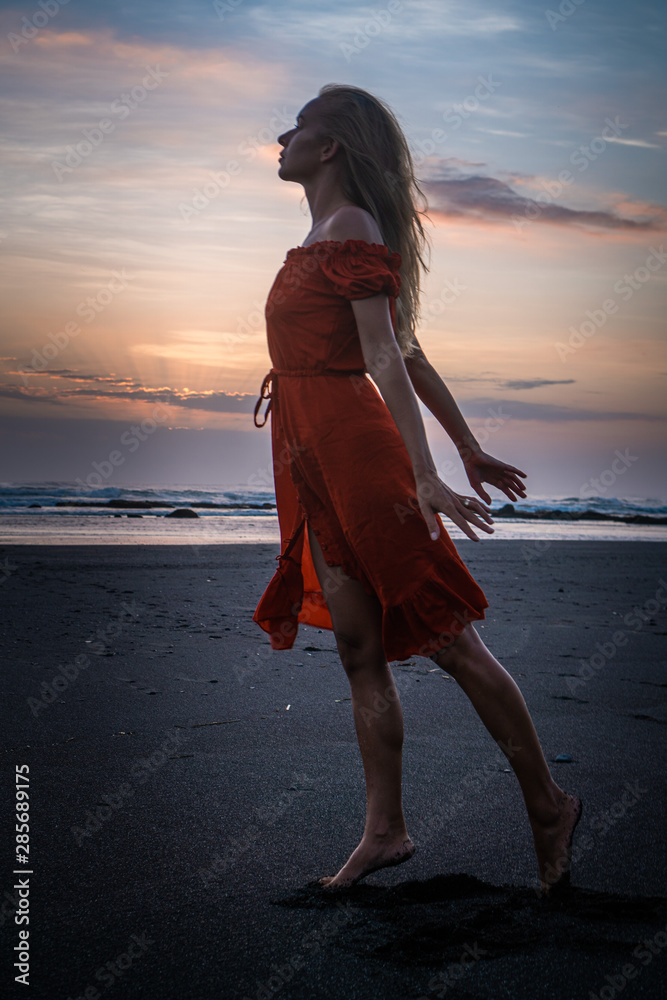 Beautiful sensual woman in red dress posing on the black sand beach during sunset