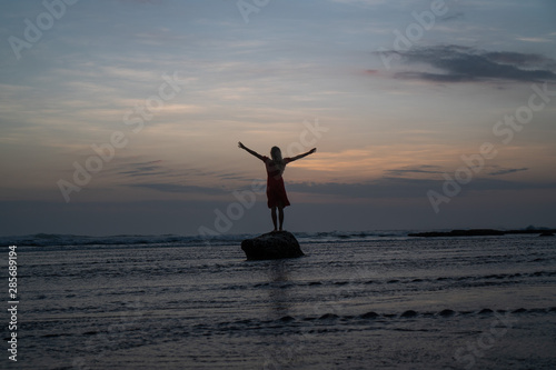 Silhouette of woman in red dress posing on the small rock in the sea water during sunset