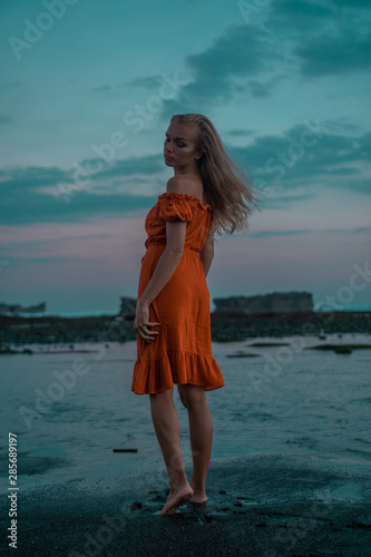 Beautiful sensual woman in orange dress posing on the black sand beach during sunset
