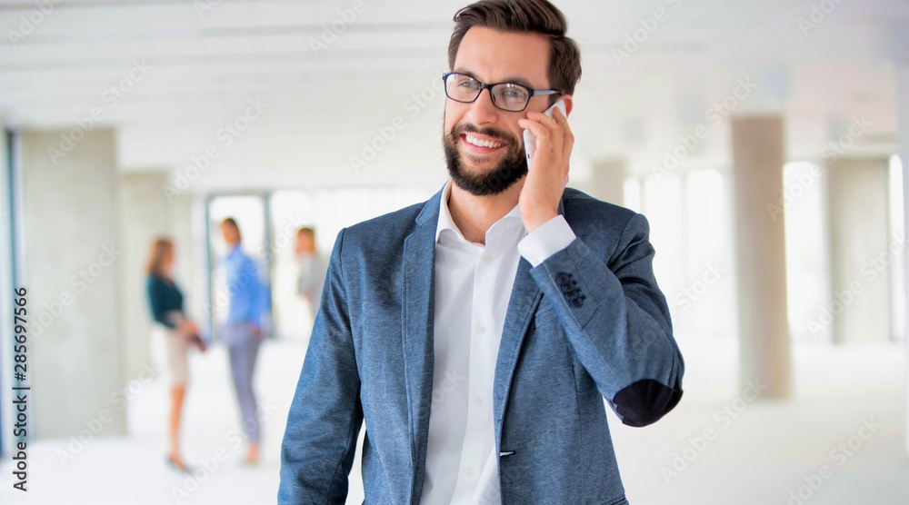 Smiling businessman wearing eyeglasses while talking on smartphone against colleagues Smiling businessman wearing eyeglasses while talking on smartphone against colleagues
