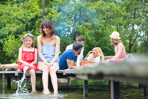 Smiling woman pointing to daughter while sitting on pier against family at lakeshore during summer © moodboard