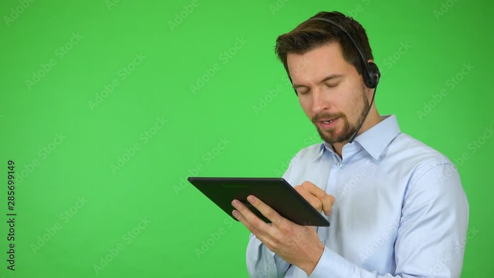 A young handsome call center agent works on a tablet and talks to a caller, then smiles at the camera - green screen studio A young handsome call center agent works on a tablet and talks to a caller, then smiles at the camera - green screen studio