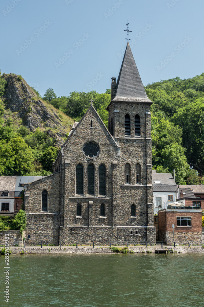 Dinant, Belgium - June 26, 2019: Gray stone Saint Paul Church among other commom dwellings along Meuse River under blue sky. Green foliage.