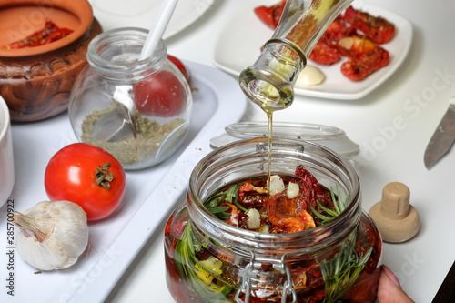 Sun-dried tomatoes with spices and garlic in a jar. Woman adds olive oil. Near rosemary, tomatoes and garlic.