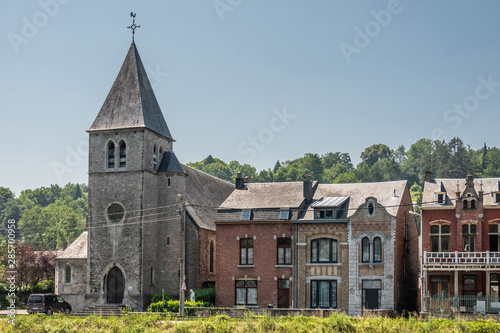 Dinant, Belgium - June 26, 2019: Gray stone Saint Paul Church among other commom dwellings along Meuse River under blue sky. Green foliage. photo