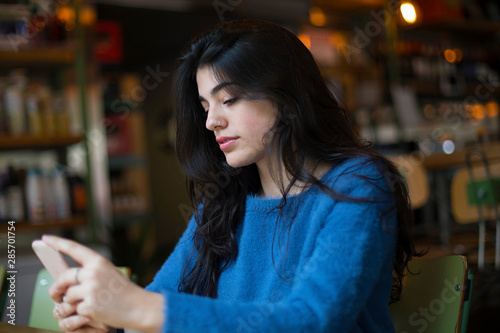 Young woman in casualwear talking through video-chat in her smartphone while sitting in cafe
