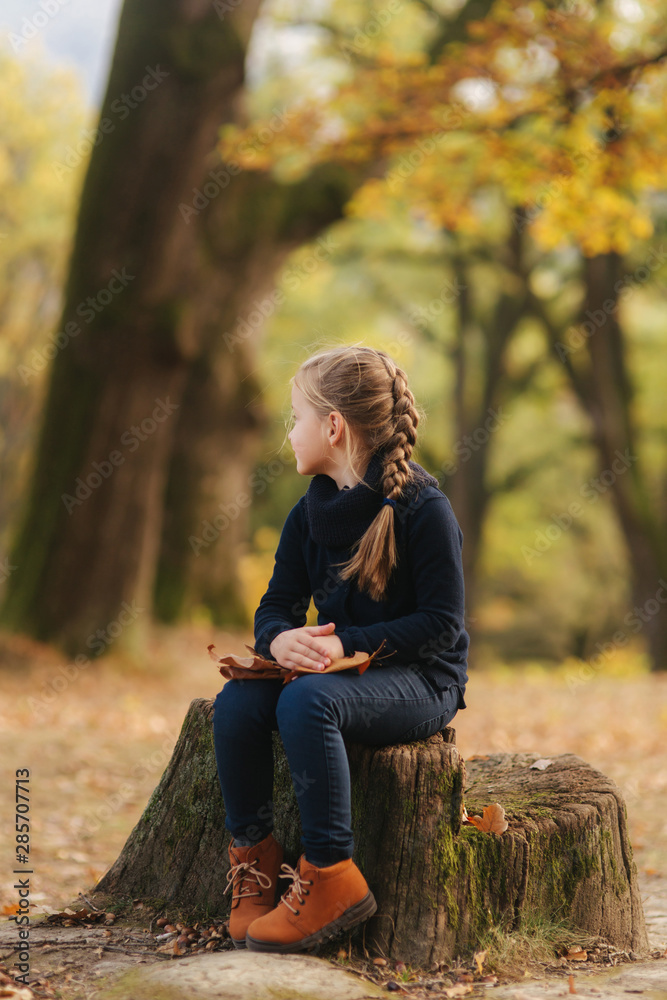 Obraz premium A little girl in black clothes is sitting on a stump and playing with autumn leaves. Blond hair girl