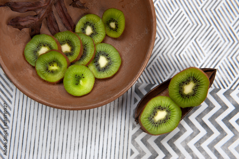 Kiwi fruit, rich in vitamin C, in a clay bowl. Pieces on top of a natural peel. Light colored background with stripes. Top view. From above. Space for text. Horizontal. Kiwi fruit, rich in vitamin C, in a clay bowl. Pieces on top of a natural peel. Light colored background with stripes. Top view. From above. Space for text. Horizontal.
