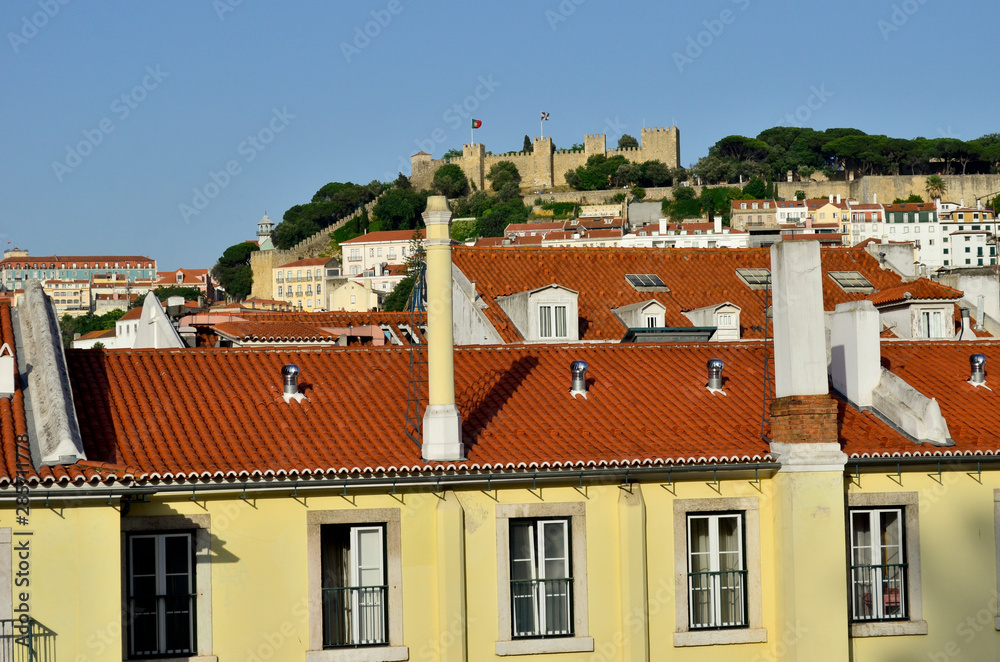 Yellow houses with red roofs with a fort in the distance behind and the blue sky, Lisboa, Portugal