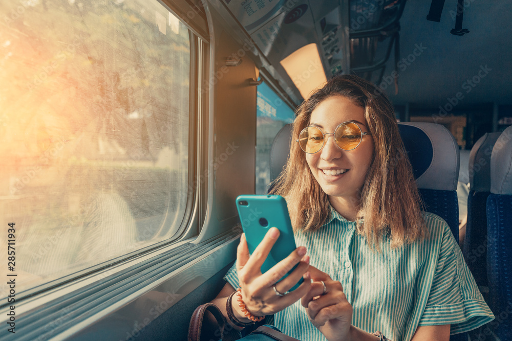 Happy Asian girl using transport application on smartphone while travelling by high speed train - obrazy, fototapety, plakaty 